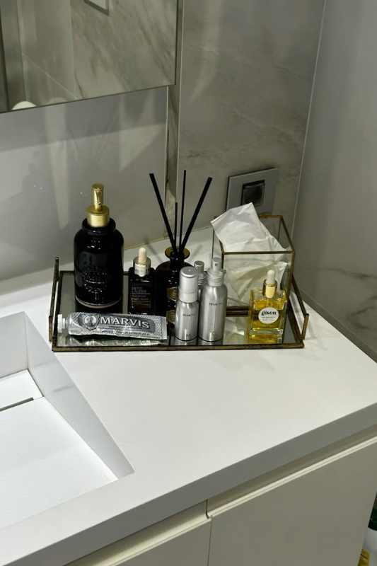 Modern bathroom vanity styled with a gold-framed glass tray holding luxury skincare products, a reed diffuser, facial oils, a tissue box, and a Marvis toothpaste tube. The clean white counter and marble wall backdrop create a sleek, organized, and upscale aesthetic. 