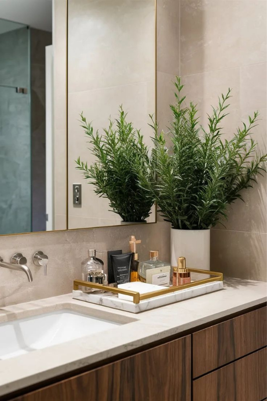 Modern bathroom counter with a marble and gold tray holding assorted skincare and perfume bottles, paired with a large potted green plant. The setup sits against a beige tiled wall with a minimalist mirror and wall-mounted faucet, creating a clean and upscale look. 