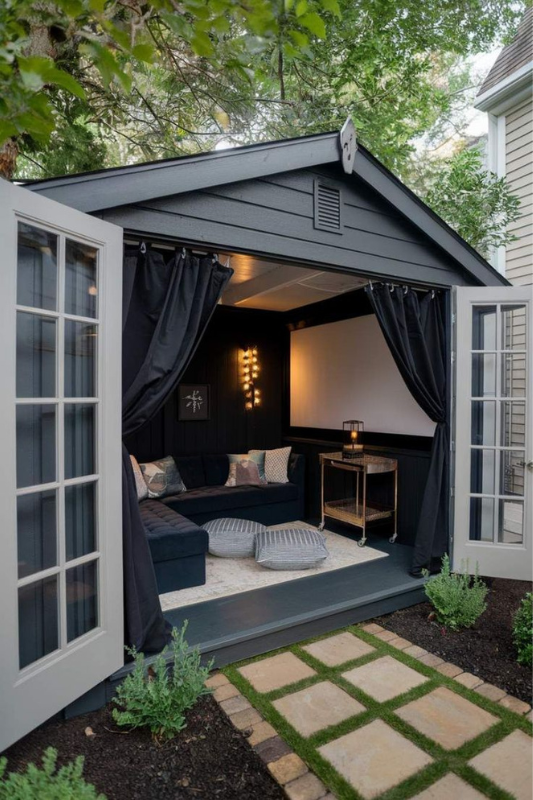 This she shed redefines backyard luxury with its bold, cinematic transformation. Painted in matte black with floor-to-ceiling blackout curtains, this setup is perfect for immersive movie nights. A plush L-shaped sectional, striped poufs, and a soft textured area rug create a relaxed lounge vibe. Add a projector screen, LED sconce lighting, and a chic bar cart for snacks and drinks, and you've got the ultimate backyard movie theater she shed that blends comfort with dramatic flair.