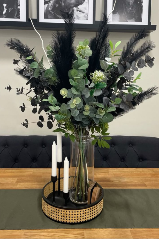 Modern dining table centerpiece featuring a tall clear glass vase filled with black pampas grass, eucalyptus, and assorted greenery, styled on a wood tray with black taper candleholders and white candles for a bold, elegant look. 