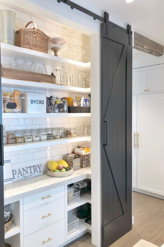 Bright and organized farmhouse pantry featuring white shelves, glass containers, wicker baskets, and a black sliding barn door.
