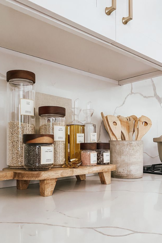 Modern kitchen counter organizer featuring glass jars with wooden lids labeled for grains and seeds, clear oil and vinegar dispensers, pink salt containers, and wooden utensils in a concrete holder, all neatly arranged on a wooden riser against a marble backsplash