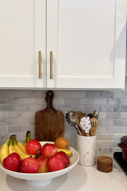 Clean kitchen counter styling with a pedestal fruit bowl filled with apples and bananas, white marble utensil holder, wooden cutting board, and gold-accented cabinet handles against a glossy subway tile backsplash