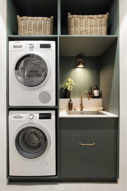 Modern compact laundry nook with stacked Bosch washer and dryer, deep green cabinetry, woven baskets, and brass faucet under warm lighting.