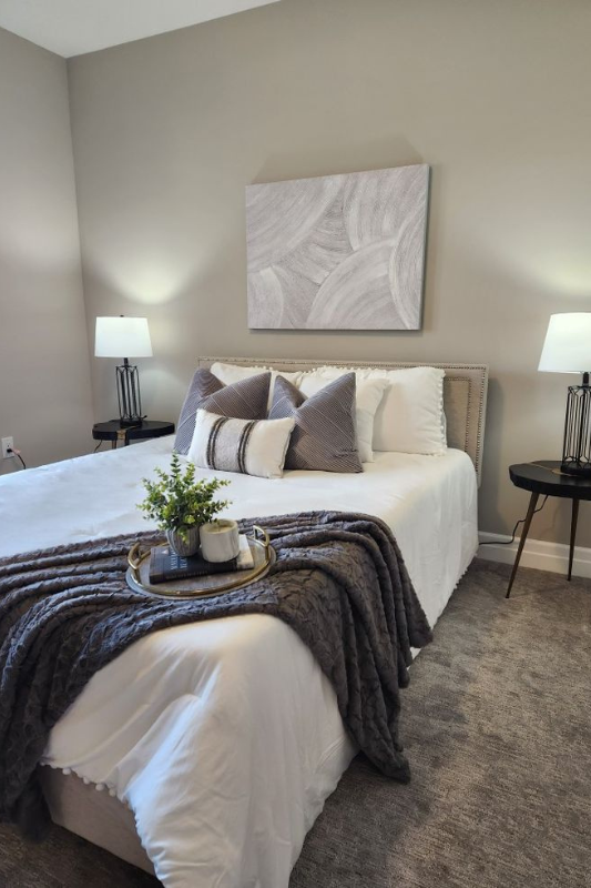 Modern neutral bedroom featuring a white bed with layered gray accent pillows, a soft textured gray throw, and a stylish round tray displaying greenery, a candle, and a coffee cup — perfect for cozy and elegant bedroom inspiration. 
