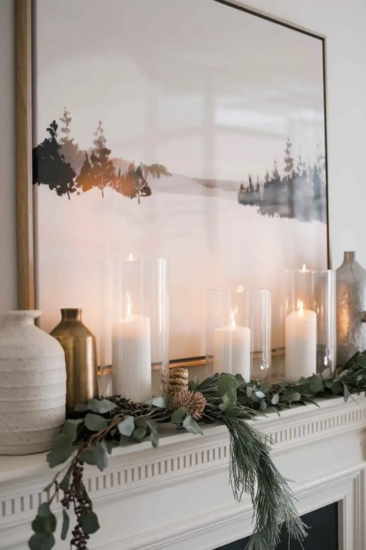 Minimalist winter fireplace mantel featuring a lush eucalyptus and pine garland accented with pinecones, paired with tall glass votives holding white pillar candles. Neutral ceramic and metallic vases complete the look beneath a serene forest landscape artwork—perfect for cozy seasonal styling, modern rustic decor, and elegant winter mantle inspiration.