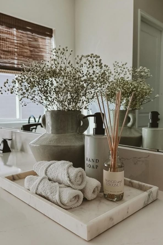 Neutral-toned bathroom counter styled with a white marble tray holding a ceramic vase of baby’s breath, a hand soap bottle, a mango and coconut reed diffuser, and rolled hand towels. The setup exudes calm, spa-like elegance with earthy textures and soft natural light. 