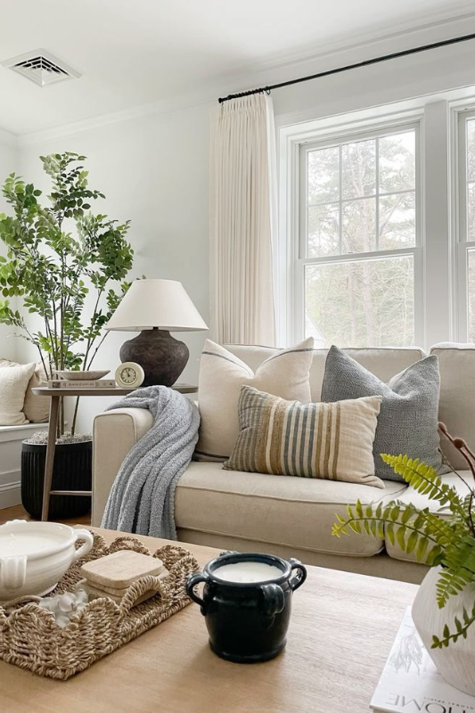Neutral coastal living room with a beige sofa styled with layered striped and solid throw pillows, textured throw blanket, ceramic lamp, large potted plant, and woven tray decor on a wooden coffee table 
