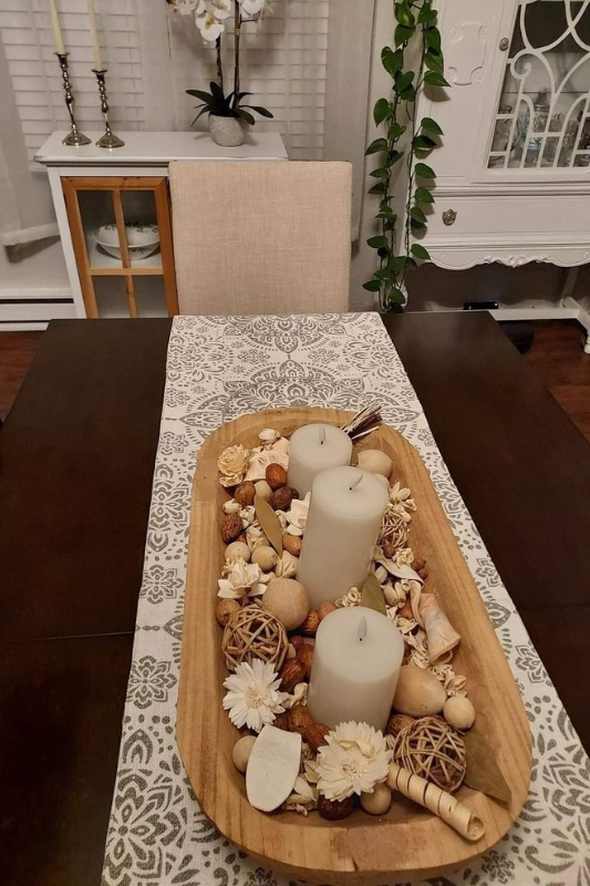 Neutral dining table centerpiece featuring a wooden dough bowl filled with cream pillar candles, decorative orbs, dried botanicals, and nuts, styled on a patterned table runner for a cozy rustic tablescape. 
