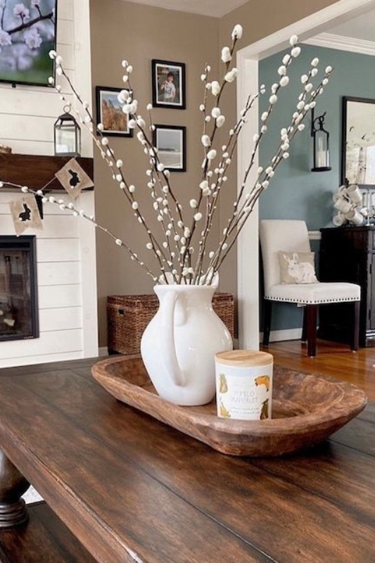 Neutral farmhouse living room with a rustic wooden dough bowl centerpiece on a dark wood coffee table, styled with a white ceramic pitcher filled with pussy willow branches and a vanilla-scented candle.
