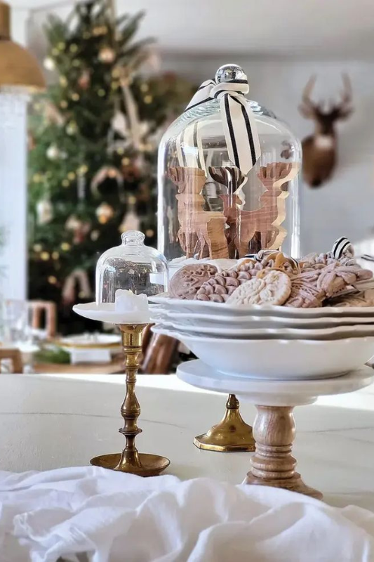Neutral holiday dining table display featuring a tiered white cake stand with decorative cookies, a glass cloche filled with wooden reindeer ornaments tied with ribbon, and brass candlestick holders, styled in a cozy cottage-inspired Christmas setting. 