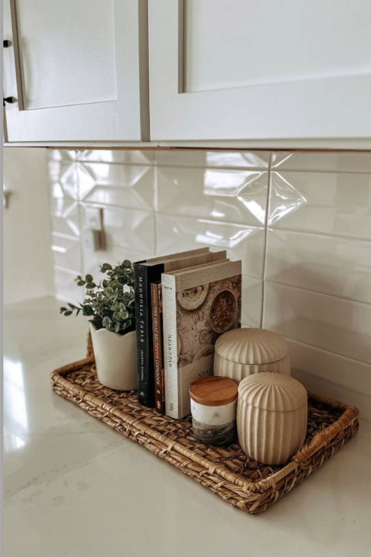 Neutral kitchen countertop decor with a woven tray holding decorative cookbooks, a small potted plant, ribbed ceramic canisters, and a marble-look backsplash for a clean and cozy aesthetic