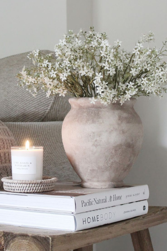 Neutral summer home decor with a rustic ceramic vase filled with white flowers, a lit lime basil and mandarin candle, and two lifestyle books styled on a wooden stool 