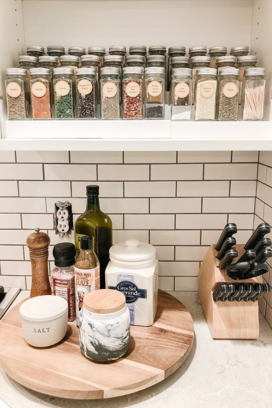 Highly organized kitchen spice shelf with labeled glass jars, paired with a round wooden tray holding salt containers, balsamic glaze, olive oil, and a pepper grinder, next to a knife block against white subway tile backsplash for a functional yet stylish setup