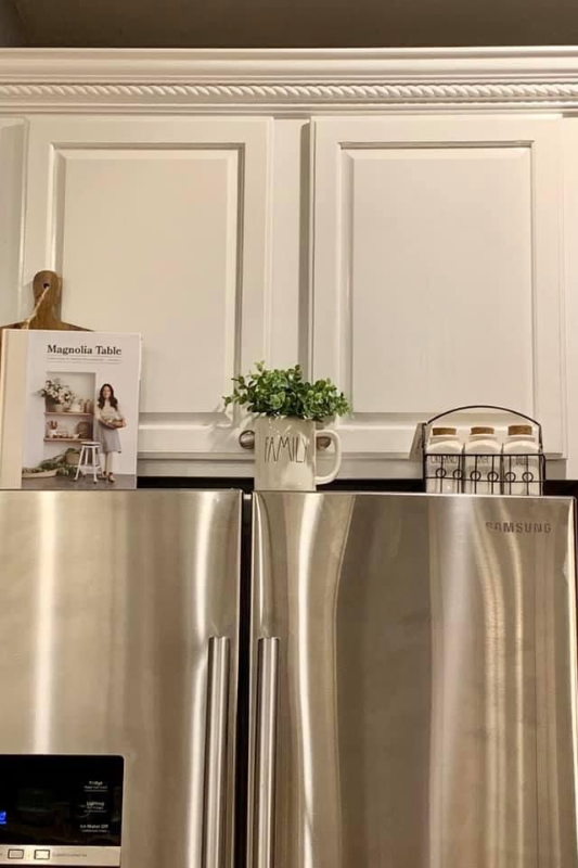 Minimalist white kitchen with Rae Dunn family mug, cookbook, and spice rack decor above stainless steel fridge