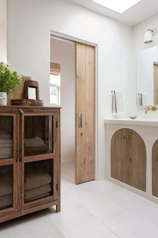 Bright bathroom space featuring a light wood pocket door, arched wood vanity cabinets, rustic cabinet with folded towels, and soft white walls with natural accents.