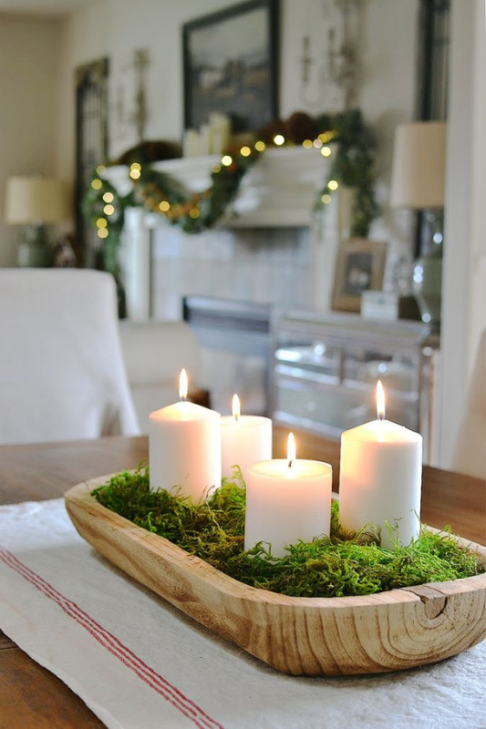 Rustic dough bowl centerpiece with white pillar candles and green moss on a farmhouse dining table with Christmas garland in the background