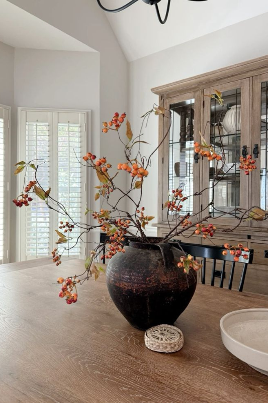 Rustic fall dining table centerpiece featuring a large black ceramic vase filled with faux orange berry branch stems, styled on a natural wood table with neutral decor for a warm and earthy seasonal look. 