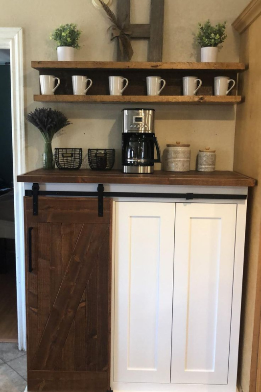 Rustic farmhouse coffee bar cabinet featuring a sliding barn door, wood countertop, upper shelf with mugs, greenery in white pots, and decorative letter “H” wall art. 