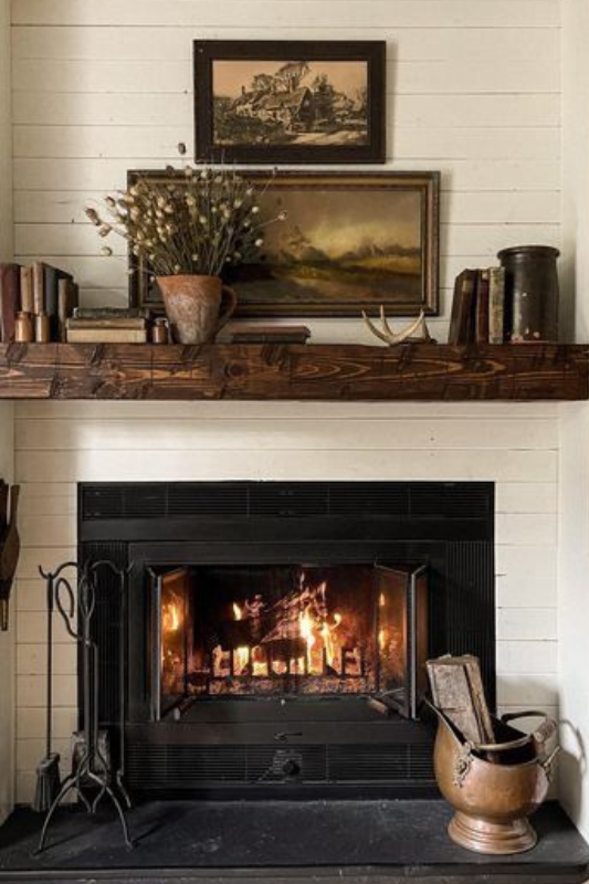 Warm rustic farmhouse fireplace featuring a dark wood mantel styled with vintage books, antique pottery, landscape paintings, and dried floral stems. The cozy shiplap wall backdrop and wood-burning fire create an inviting, old-world charm—perfect for cottagecore interiors, traditional fall decor, and cozy vintage-inspired living room aesthetics.