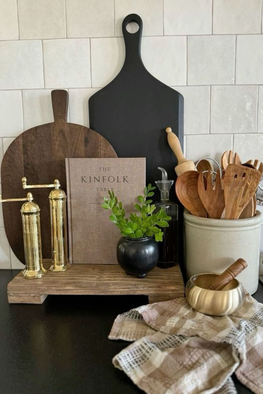 Rustic kitchen counter vignette featuring wooden cutting boards, brass salt and pepper grinders, ceramic utensil holder, mini black planter with greenery, and a linen towel on a matte black countertop with light tile backsplash