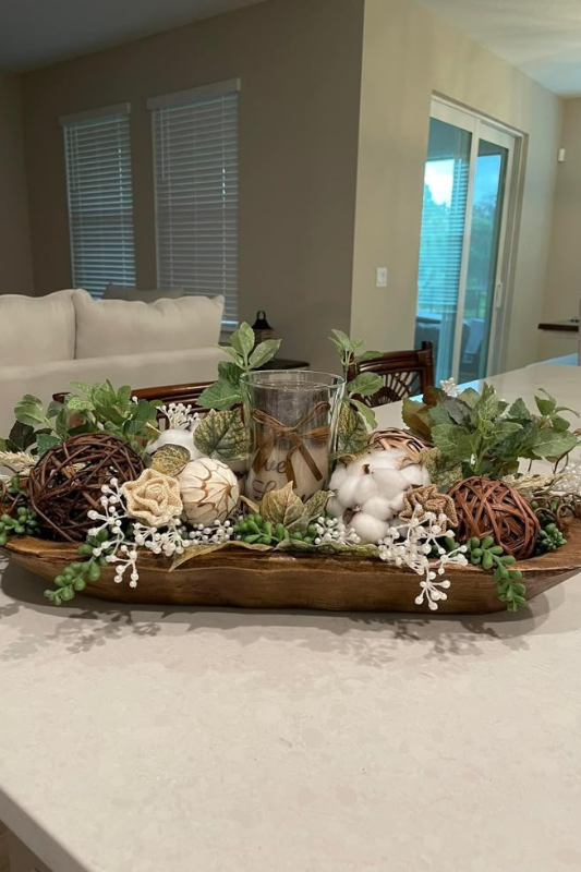 Decorative spring dough bowl centerpiece on a white kitchen island, filled with green foliage, cotton stems, rattan balls, artificial florals, and a rustic candle in a glass holder wrapped with twine.