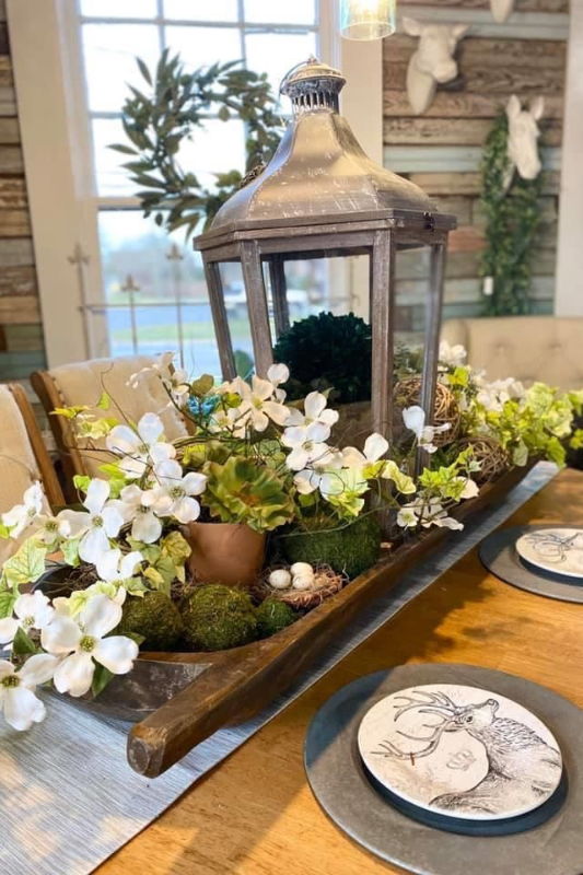 Charming spring dough bowl centerpiece on a dining table featuring a tall rustic lantern surrounded by white faux flowers, moss balls, greenery, a small terracotta pot, and decorative bird eggs for a fresh farmhouse look.