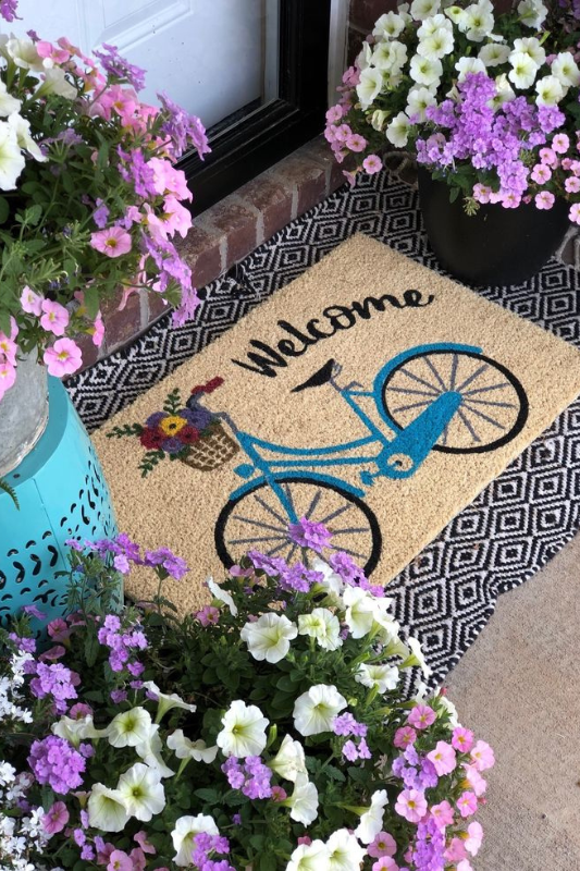 Charming summer front porch with a welcome mat featuring a blue bicycle and flower basket, surrounded by vibrant pink and white petunias, layered over a black and white geometric outdoor rug for a cheerful seasonal entryway display 