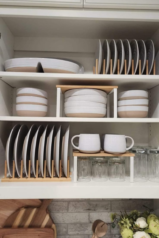 Modern white kitchen cabinet organized with vertical bamboo dish racks, showcasing neatly stacked dinnerware, bowls, mugs, and glassware for smart cabinet organization. 