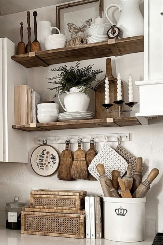Beautiful vintage farmhouse kitchen decor featuring open wooden shelves with white ceramic pitchers, plates, greenery, framed butterfly art, and taper candles. Below, a counter is styled with woven baskets, books, a ceramic utensil crock with rolling pins, hanging brushes, and knitted pot holders, creating a warm and charming rustic aesthetic.