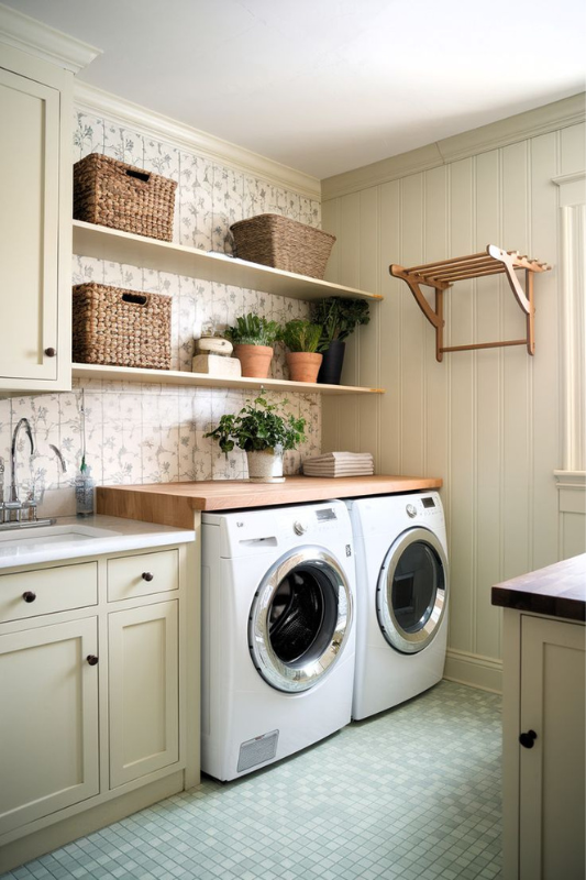Charming farmhouse laundry room with floral wallpaper, open shelves, potted plants, and wood countertop above front-load machines.