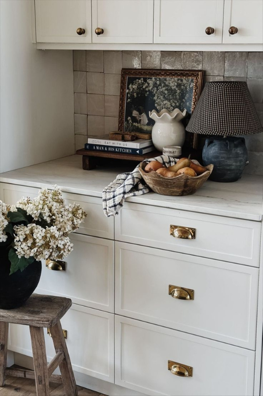 Vintage-inspired kitchen counter styling featuring cream cabinetry with brass cup pulls, a framed floral art print, stacked cookbooks, a wooden fruit bowl with pears, a gingham-shaded ceramic lamp, and a vase of white hydrangeas on a rustic wooden stool against a taupe tiled backsplash