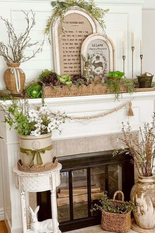 Whimsical vintage spring fireplace styled with botanical-themed art, a rustic wooden bead garland, and greenery spilling from the mantel. Natural textures like moss, wicker, and terra cotta pots complement potted florals, taper candles, and antique-style vessels. A Velveteen Rabbit quote and French rabbit print add storybook charm—perfect for spring cottage decor, farmhouse living rooms, and garden-inspired mantel styling.