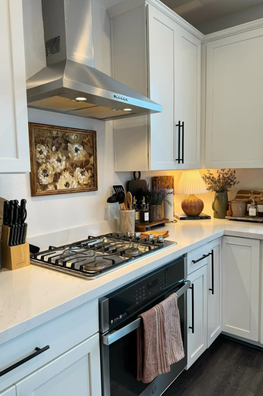Warm modern kitchen decor featuring white shaker cabinets, a stainless steel range hood, floral wall art, and cozy countertop styling with a ceramic lamp, greenery in a vase, oil and spice containers, and a coffee station setup in the corner for a functional yet inviting cooking space