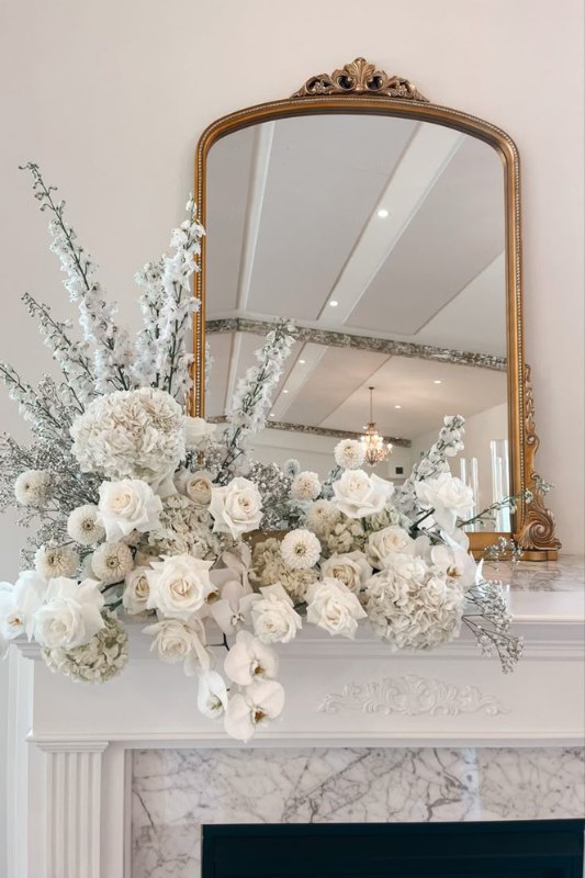 Luxurious white floral arrangement on a classic white fireplace mantle, showcasing premium roses, hydrangeas, orchids, and baby’s breath. Styled with a gold ornate vintage mirror and elegant marble detailing, perfect for upscale home decor, wedding inspiration, or spring mantle styling ideas.