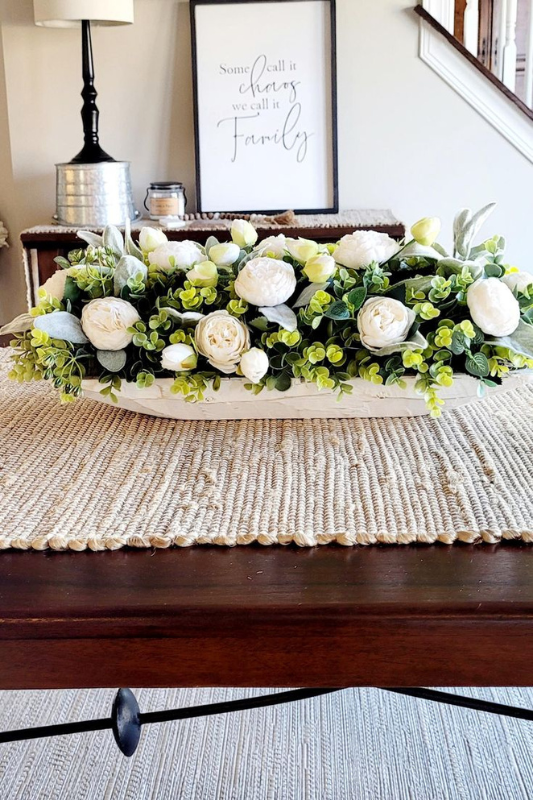 Elegant farmhouse dough bowl centerpiece filled with white roses, peonies, and soft green foliage, placed on a woven table runner atop a dark wood dining table in a cozy, family-themed living space.