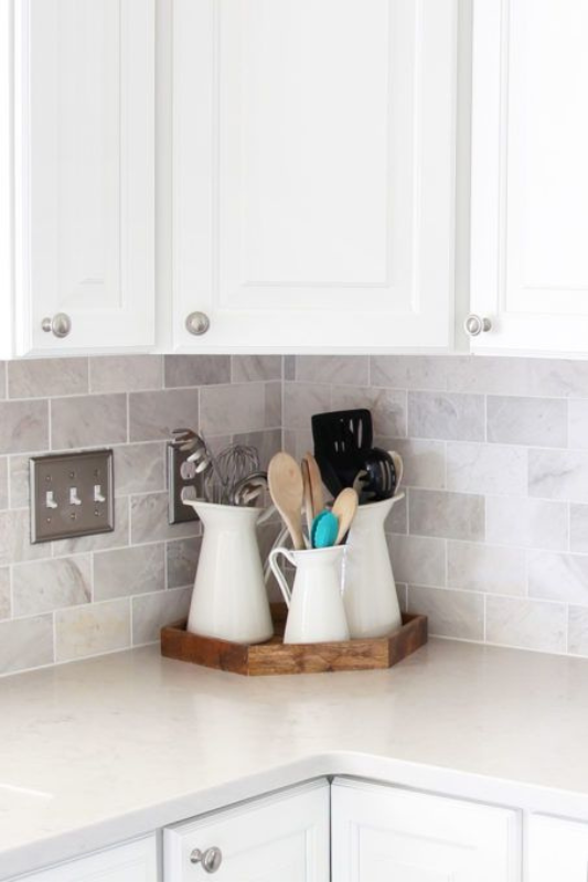 Clean white kitchen corner styled with white ceramic pitchers holding wooden and metal utensils, all arranged on a wooden tray atop a neutral quartz countertop and subway tile backsplash for a simple, organized look