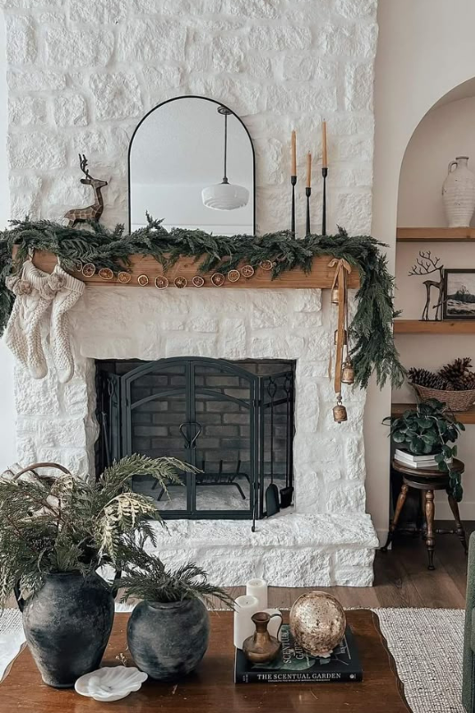Cozy white stone fireplace decorated with a lush evergreen garland, dried orange slices, gold bells, and black taper candles in sleek holders. A minimalist arched mirror and festive reindeer figurine sit atop the wood mantel, blending rustic charm with modern holiday styling. The space is accented with natural greenery, earthy vases, and a wood coffee table—perfect for winter decor ideas, modern farmhouse living rooms, and Christmas mantel inspiration.