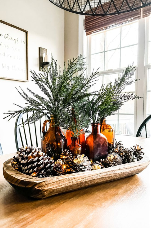Rustic winter dough bowl centerpiece displayed on a wooden dining table, featuring amber glass bottles filled with evergreen stems, surrounded by pinecones, dried florals, and warm string lights for a cozy seasonal touch.