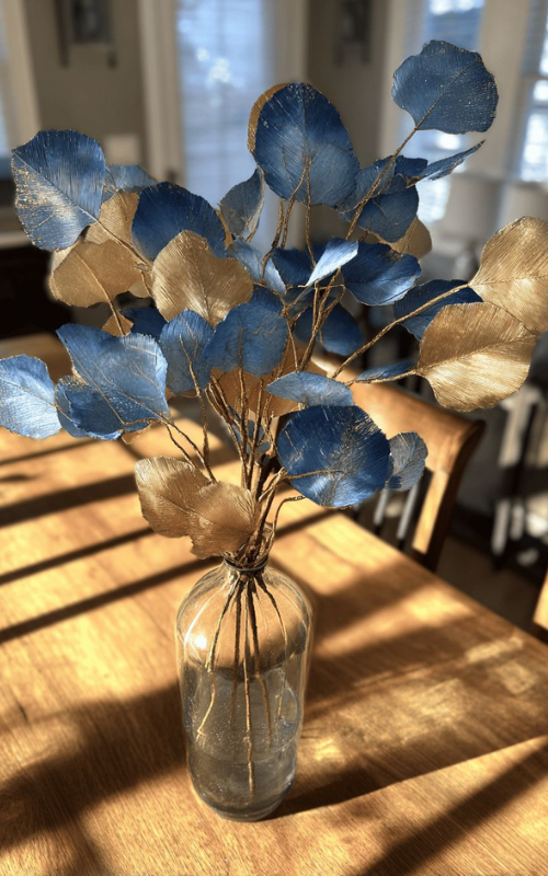 Blue and gold painted dried leaves in a clear glass vase on a wooden fall dining table.