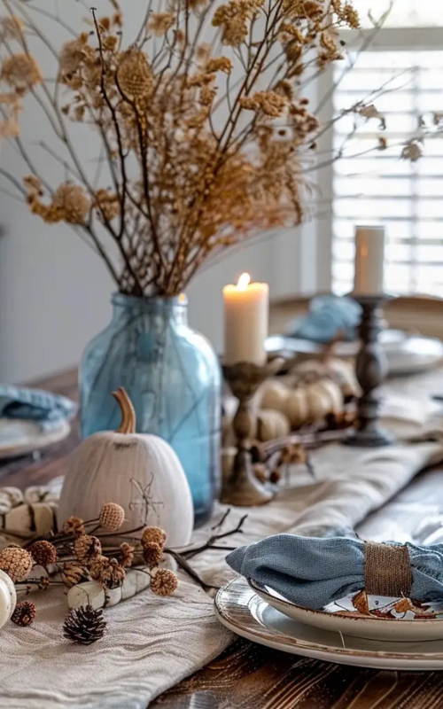 Fall table with blue glass vase, white pumpkins, rustic candles, and dried orange flowers for an elegant boho look.