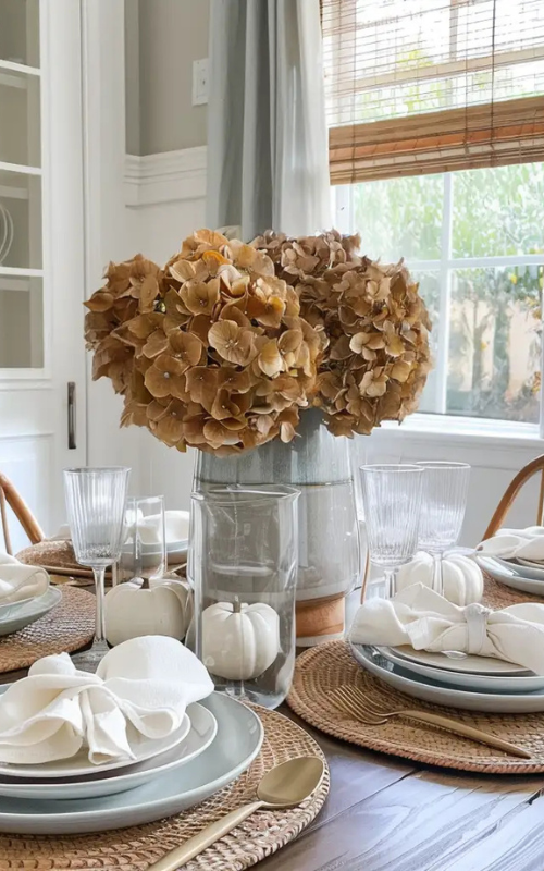 Elegant fall table with blue-gray plates, gold cutlery, white pumpkins, and neutral hydrangeas in a glass vase.